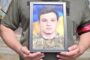 Person in a green military uniform holding a framed photo of a young man in camouflage.