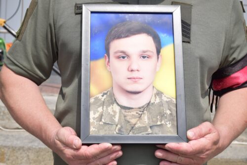Person in a green military uniform holding a framed photo of a young man in camouflage.