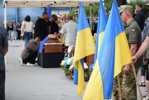 Mourners gather for a funeral under a canopy, with a casket draped in black and blue and Ukrainian flags in the foreground.