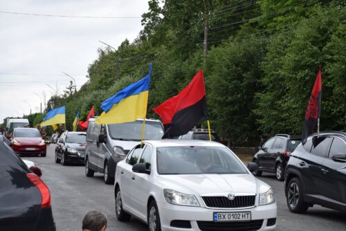 Car convoy in a tree-lined street displaying Ukrainian blue-yellow flags and red-black flags from vehicles