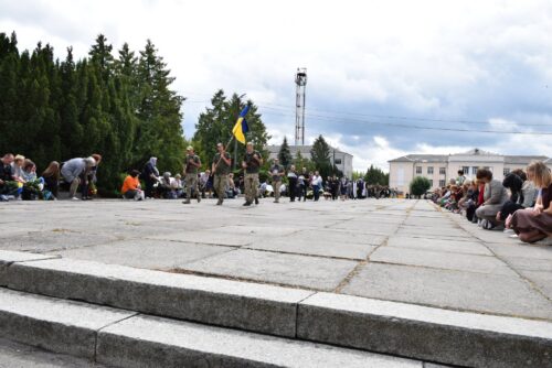 Public ceremony on a wide paved square with soldiers carrying a Ukrainian flag; spectators sit along the edges of the square.