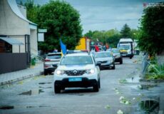 Convoy of cars in a street parade with blue and yellow flags, led by a white Renault SUV on a wet, puddled road.