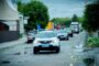 Convoy of cars in a street parade with blue and yellow flags, led by a white Renault SUV on a wet, puddled road.