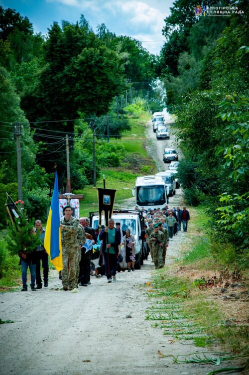 Funeral procession on a rural dirt road, led by a man holding the Ukrainian flag and a cross, followed by civilians, soldiers, and a line of cars.