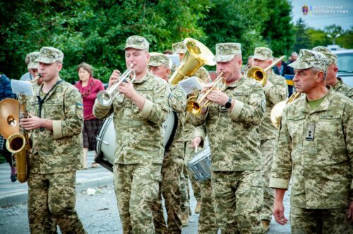 Military band in camouflage uniforms playing brass instruments outdoors during a public event, with spectators in the background.