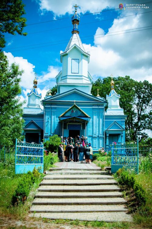 Blue wooden church with onion domes and white trim, people gather at the entrance on stone steps on a sunny day.