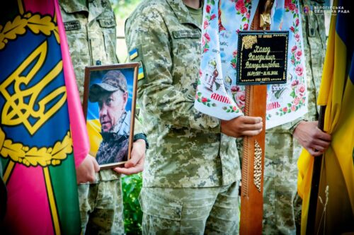 Ukrainian soldiers in camouflage hold a framed portrait and a carved cross during a memorial ceremony with flags nearby.