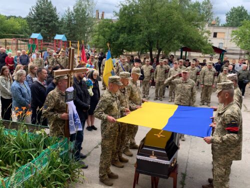 Military funeral ceremony: soldiers present a coffin draped with a blue and yellow Ukrainian flag while civilians and a priest hold a cross nearby.
