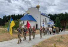Soldiers in camouflage with sashes march past a small white church with a blue roof and golden domes, carrying the Ukrainian flag and another ceremonial banner.