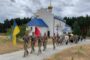 Soldiers in camouflage with sashes march past a small white church with a blue roof and golden domes, carrying the Ukrainian flag and another ceremonial banner.