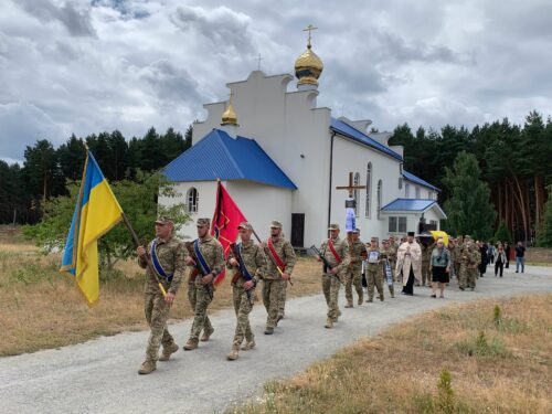 Soldiers in camouflage with sashes march past a small white church with a blue roof and golden domes, carrying the Ukrainian flag and another ceremonial banner.