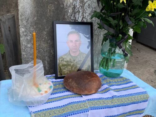 A makeshift memorial altar with a framed portrait of a man in a military uniform, a lit candle, bread, a drink, and flowers on a patterned cloth.