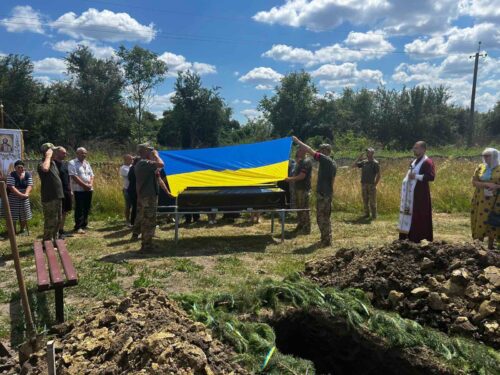 Mourners surround a coffin draped with a blue-and-yellow Ukrainian flag at a gravesite in a grassy field, with a priest and onlookers nearby.