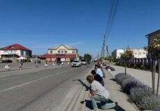 Sunny small-town street with spectators crouching by the curb to watch an event, buildings across the road.