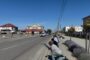 Sunny small-town street with spectators crouching by the curb to watch an event, buildings across the road.