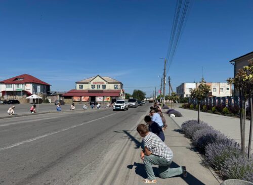 Sunny small-town street with spectators crouching by the curb to watch an event, buildings across the road.
