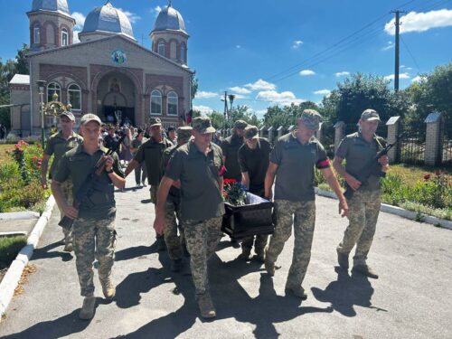 Military pallbearers in camouflage carry a flag-draped coffin during a funeral procession past a brick church on a sunny day.