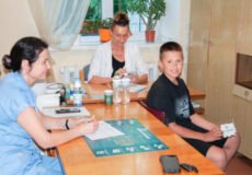 Two adults, likely clinic staff, sit at a desk with forms and bottles while a smiling boy patient sits nearby holding cards in a treatment room.