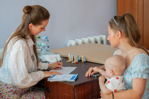 Healthcare professional sits at a desk reviewing documents with a mother and her baby present at a clinic table with supplies nearby.