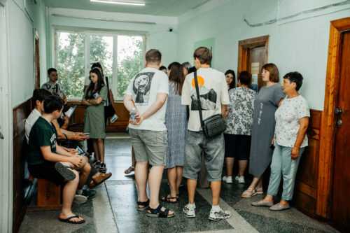 Group of young people standing and sitting in a pale-green hallway, waiting near a wooden door.