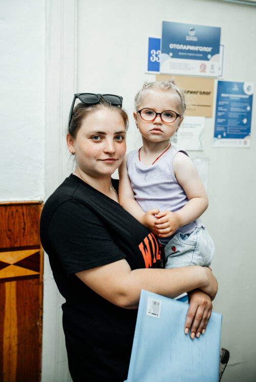 Mother holding a toddler with glasses in a clinic hallway, posters on the wall behind them.