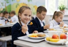 Schoolchildren eating lunch in a bright classroom; girl in the foreground smiles at the camera with a fork and plate of food ahead.