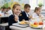 Schoolchildren eating lunch in a bright classroom; girl in the foreground smiles at the camera with a fork and plate of food ahead.