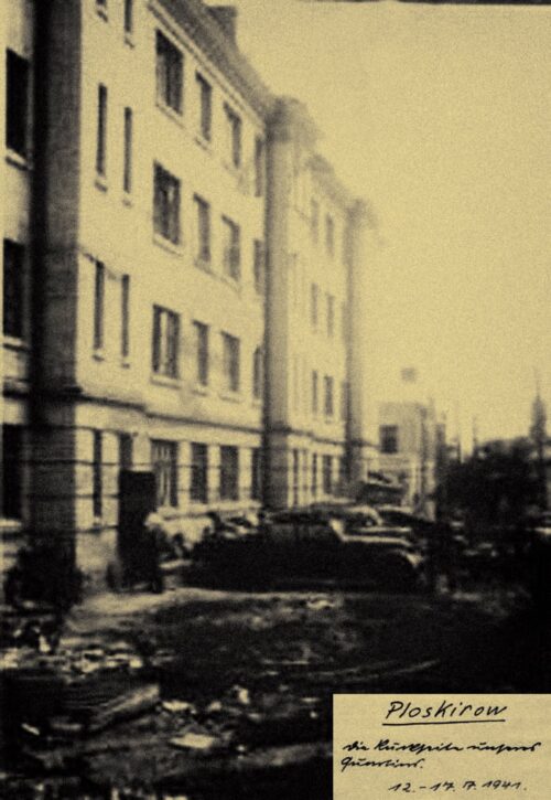 Historic street scene with a row of apartment buildings, debris on the road, and vintage cars; a handwritten note in the bottom-right corner reading Ploskirow.