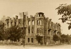 Sepia-toned photo of a ruined three-story stone building with arched windows on a tree-lined street.