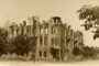 Sepia-toned photo of a ruined three-story stone building with arched windows on a tree-lined street.