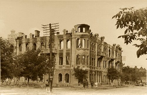 Sepia-toned photo of a ruined three-story stone building with arched windows on a tree-lined street.