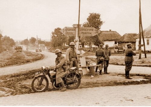 Historical WWII checkpoint with soldiers and a parked motorcycle at a dirt crossroads in a village.