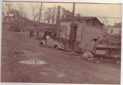 Old street in ruins with damaged cars and rubble, including a truck and debris in a desolate, wartime setting.