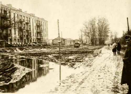 Winter street scene with a muddy, puddled road, snow on the sidewalk, and people walking beside row houses.