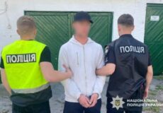 A handcuffed man in a light gray hoodie is escorted by two police officers, one in a neon vest and one in a dark uniform with Ukrainian police insignia, in front of green doors.