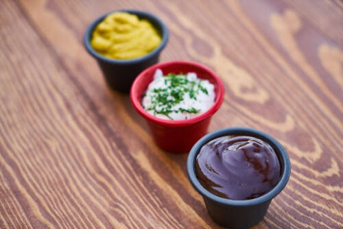 Three small condiment cups on a wooden table: mustard, sour cream with chives, and barbecue sauce (from back to front).