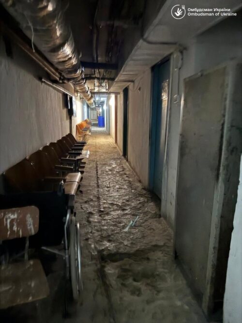 Dim concrete, dimly lit hallway with exposed pipes above; brown chairs lined along the left wall and a wheelchair at the foreground, blue door at the far end.
