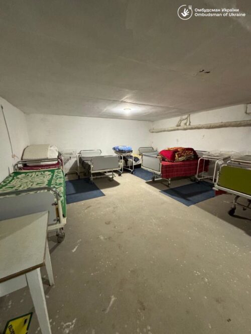 Underground shelter room with several metal hospital beds arranged in rows, some with blankets, a bare concrete floor and dim overhead lighting.