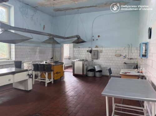Dilapidated, empty kitchen with tile walls, metal tables, and old equipment, plus exposed ceiling ducts and pipes.