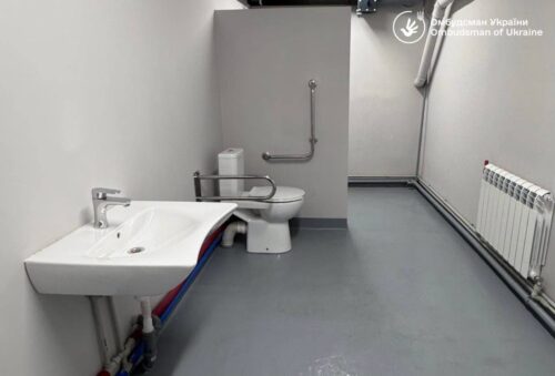 Accessible restroom with open floor space, a wall-mounted sink with exposed plumbing, a toilet equipped with grab bars and a vertical support rail, and a radiator along the right wall.