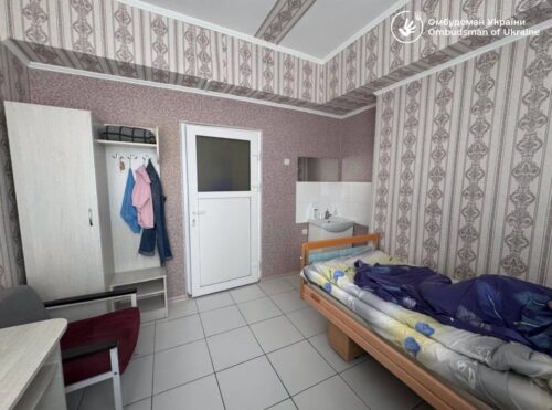 Small furnished room with a single bed, wardrobe with clothes, a desk and red chair, tiled floor, and a door.
