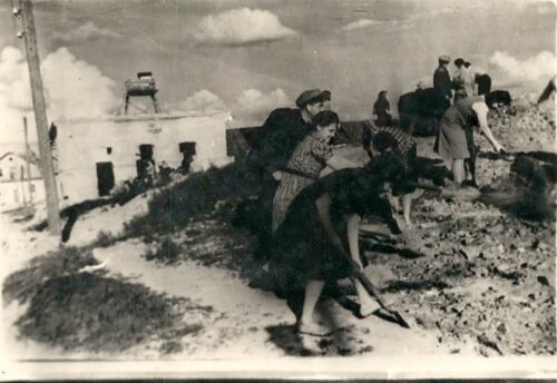 Black-and-white historical photo showing several women and children digging or shoveling on a sandy dune near a small coastal village.