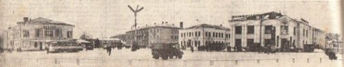 Historic town square with low-rise buildings and a tall central monument in the street era.