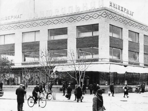 Historic black-and-white photo of a department store on a city square, with pedestrians and cyclists in front.