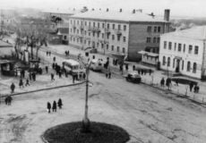 Historic black-and-white photo of a town square with trams, cars, and pedestrians around a central circular island.