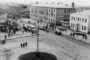 Historic black-and-white photo of a town square with trams, cars, and pedestrians around a central circular island.