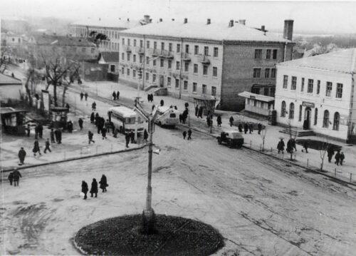 Historic black-and-white photo of a town square with trams, cars, and pedestrians around a central circular island.