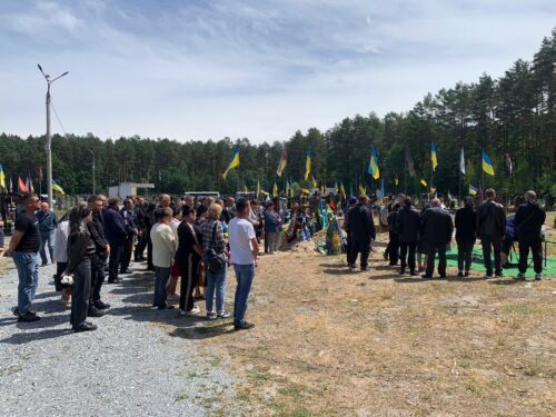 People standing in two groups at an outdoor ceremony with yellow and blue Ukrainian flags along a gravel path and a forest backdrop.