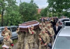 Military funeral procession with uniformed soldiers carrying a dark wooden coffin draped in a cloth, flanked by civilians and parked cars on a tree-lined street.