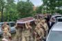 Military funeral procession with uniformed soldiers carrying a dark wooden coffin draped in a cloth, flanked by civilians and parked cars on a tree-lined street.